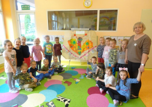 Grupa dzieci wraz z panią Agnieszką stoi wokół tablicy z przywieszonym plakatem.